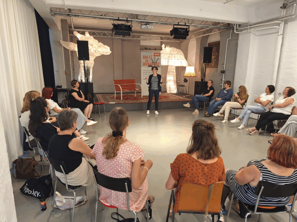 Katrin spricht vor einer Runde von rund 20 Frauen über Einfache Sprache. Ort ist das Kapuziner Kreativzentrum in Ravenburg. Im Hintergrund sind Lichtskulpturen vom Lichterfest zu sehen – ein Giraffenkopf und ein Leuchtpilz. Die Atmosphäre ist locker entspannt.