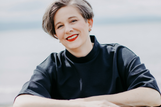 Katrin steht an einer Mauer. Zu sehen ist nur der Oberkörper. Sie lacht in die Kamera und hat die Arme auf der Mauer verschränkt.