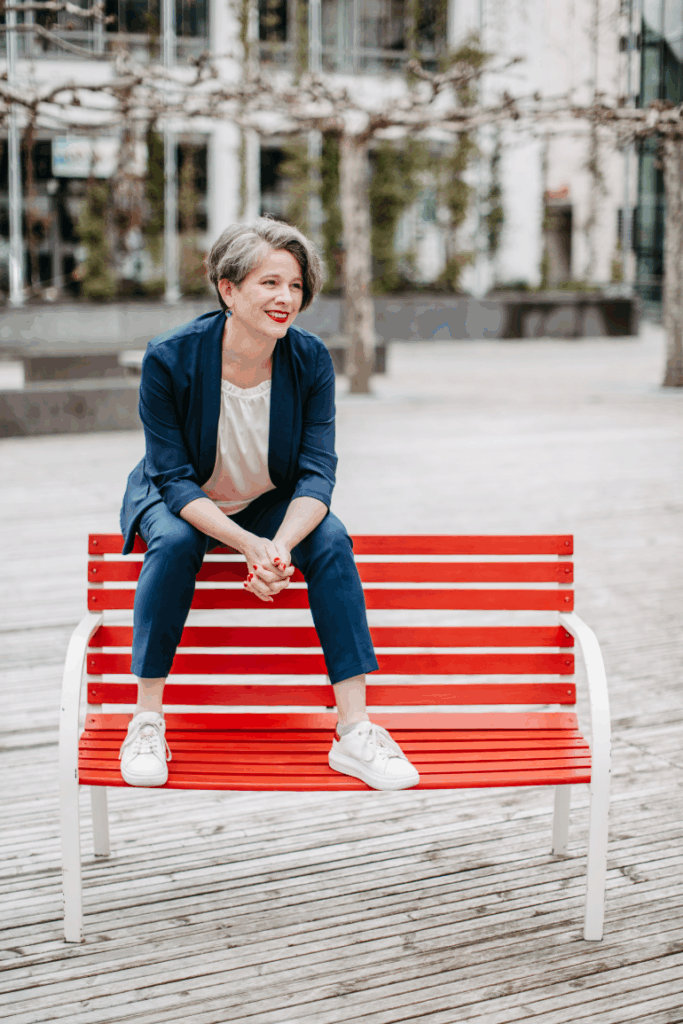 Katrin sitzt auf der Rückenlehne der roten Satzbank. Die Armlehnen sind weiß gestrichen. Ihre Füße stehen auf der Sitzfläche, sie lehnt sich leicht vor und schaut zur Seite. Katrin trägt weiße Turnschuhe und eine weiße Hose. Jacke und Hose sind dunkelblau. Im Hintergrund sind unscharf Bäume und Gebäude zu erkennen.