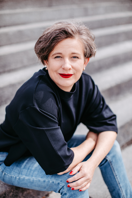 Katrin sitzt auf einer Steintreppe. Sie lacht in die Kamera. Die Arme hat sie auf ihren Knien verschränkt.