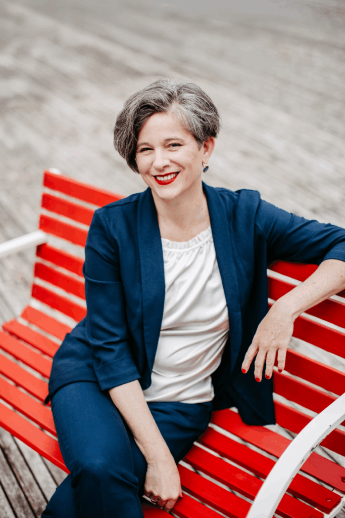 Katrin sitzt auf der roten Satzbank. Sie hat die Beine überschlagen und trägt eine weiße Bluse und eine dunkelblaue Hose und Blazer.
