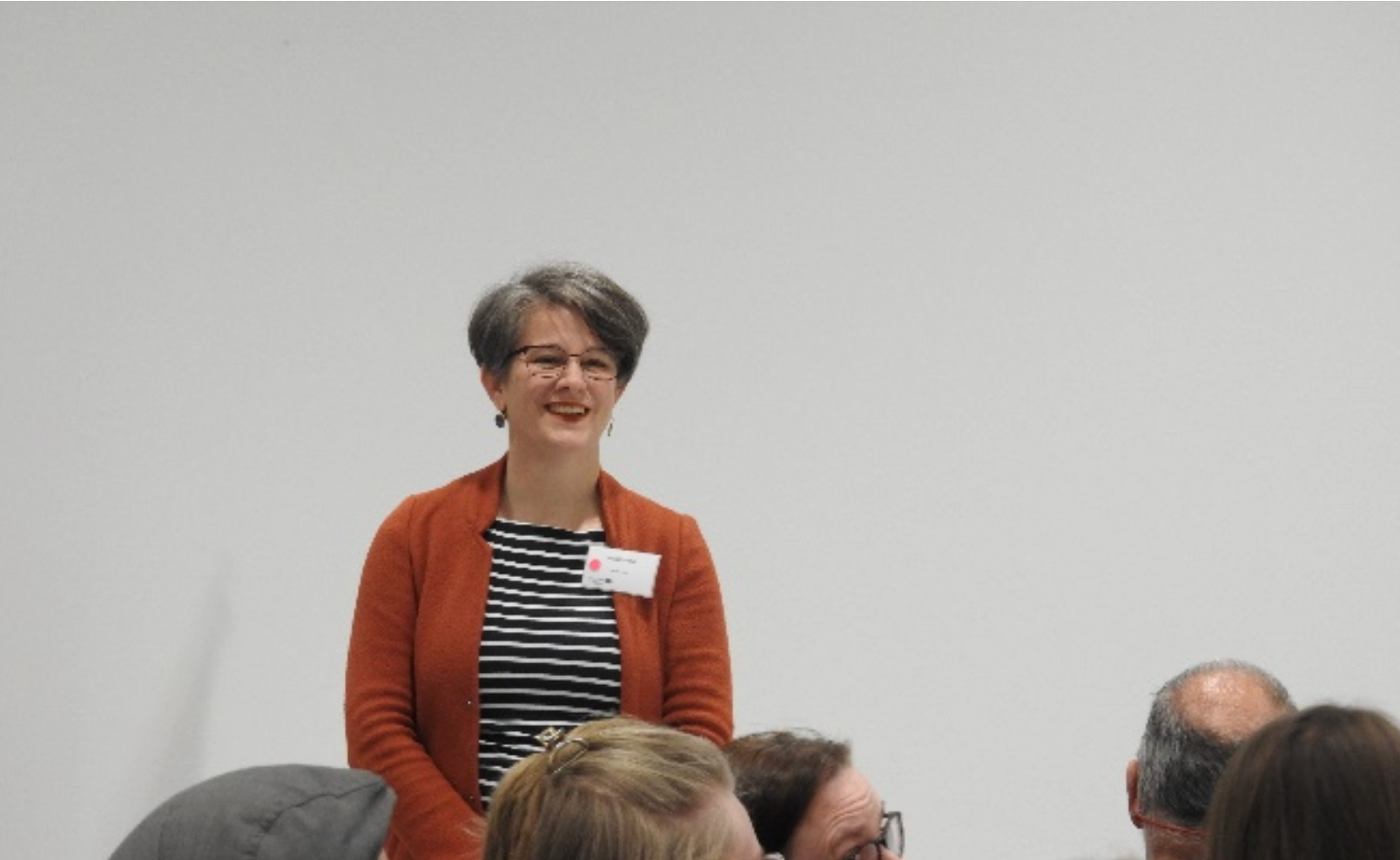 Katrin steht vorne vor einem Publikum und lacht. Sie trägt ein blau-weiß-gestreiftes T-shirt und eine orangene Jacke. Die Köpfe der Zuschauenden sind nur angeschnitten erkennbar.